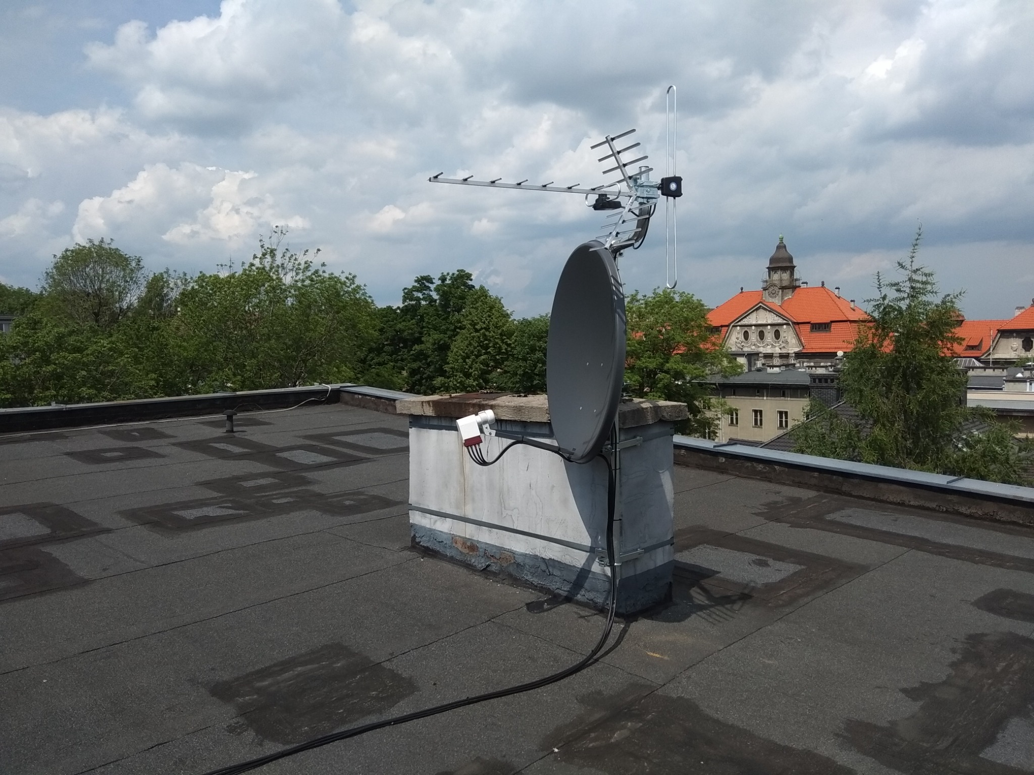 Antena satelitarna i naziemna zamontowana na kominie na dachu budynku. W tle widoczne budynki i drzewa pod pochmurnym niebem.