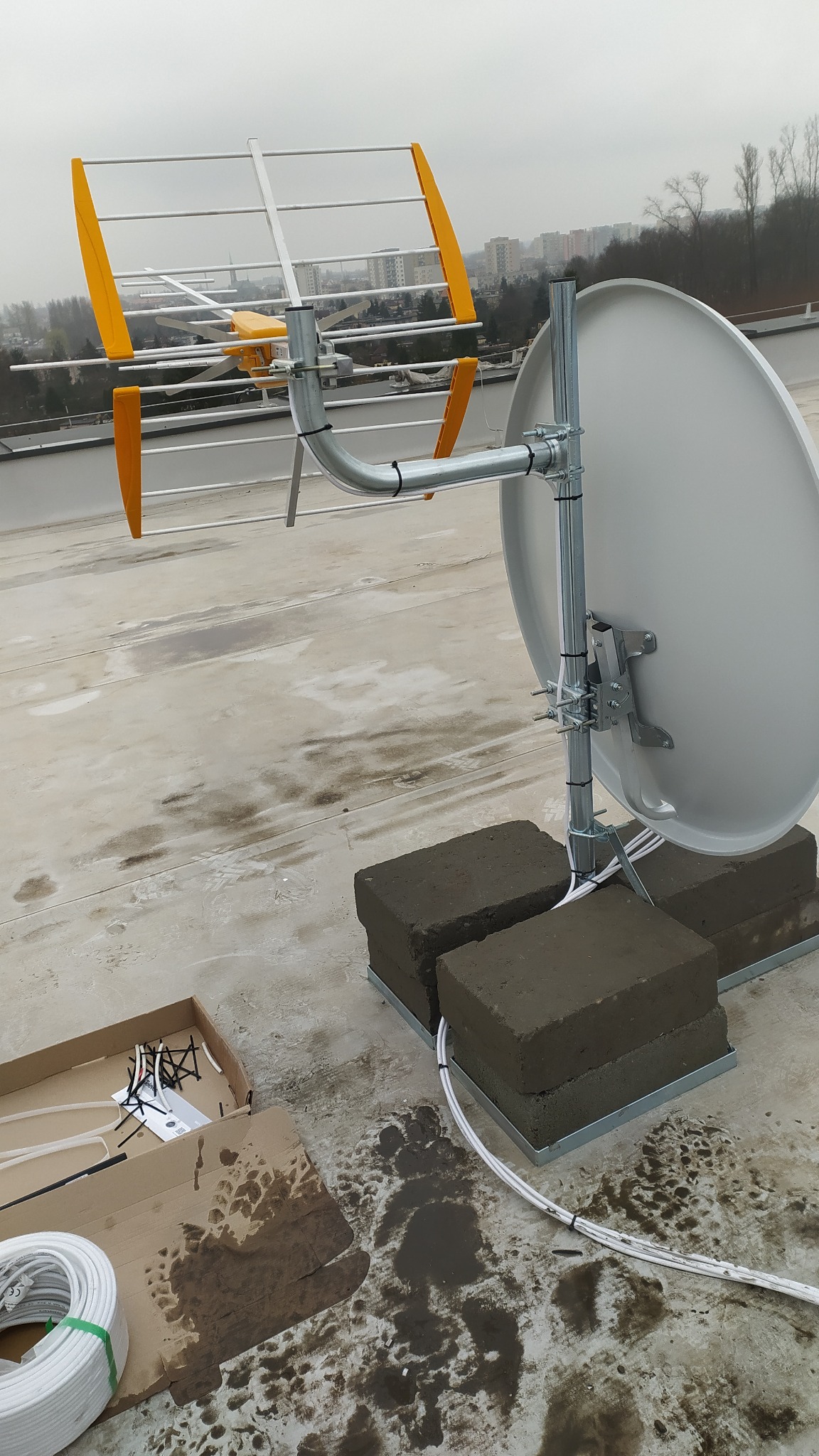 Antena satelitarna i naziemna zamontowane na dachu budynku, widoczne kable i elementy montażowe. W tle zabudowa miejska i pochmurne niebo.