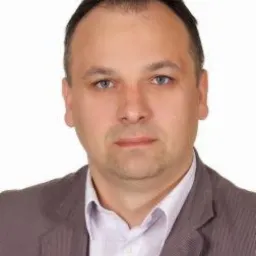 Portret mężczyzny w szarej marynarce na białym tle. Widoczne delikatne cienie podkreślające rysy twarzy. Ujęcie od przodu, skupione na twarzy i górnej części tułowia.