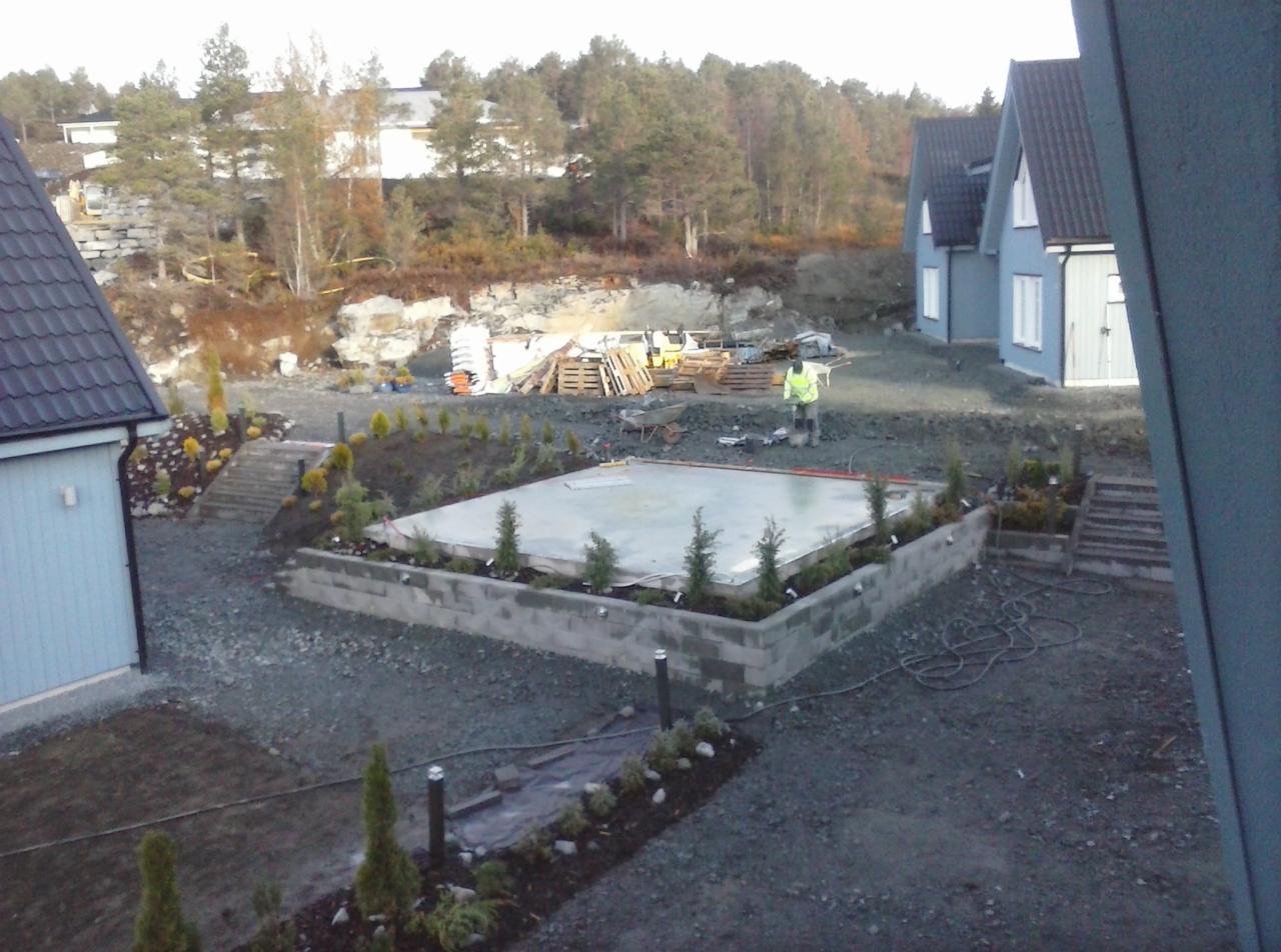 Przygotowanie terenu pod instalacje teletechniczne: wyrównany teren z betonową wylewką otoczoną niskim murem z bloczków, świeżo posadzone iglaki, widoczny pracownik w kamizelce odblaskowej...