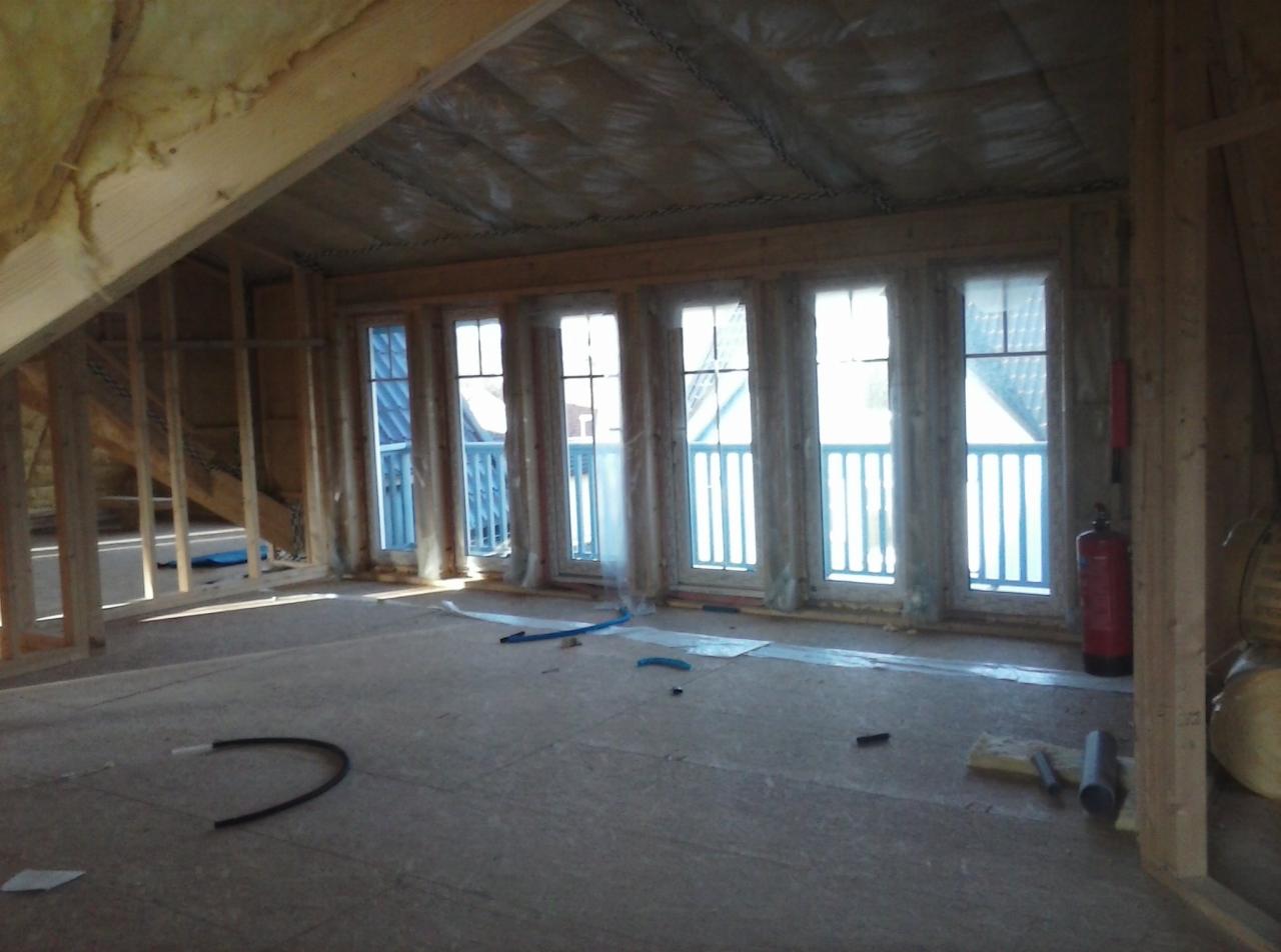 Poddasze w trakcie adaptacji w Gdańsku, widoczne drewniane elementy konstrukcyjne, ocieplenie wełną mineralną, okna balkonowe z niebieskimi balustradami na tle jasnego światła dziennego,...