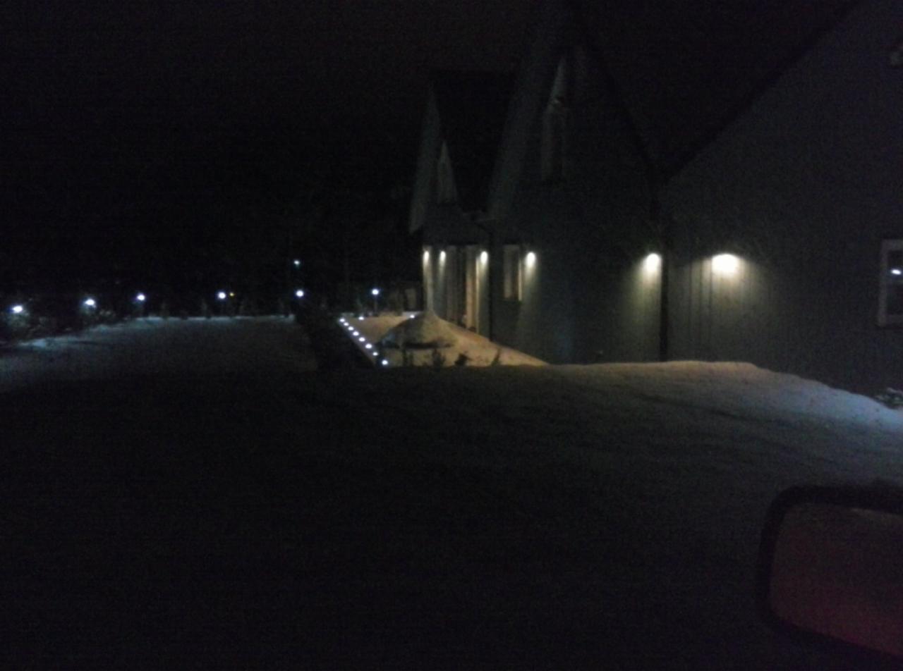 Nocne oświetlenie elewacji budynku z dwuspadowym dachem i ścieżki prowadzącej do wejścia, z widocznymi lampami gruntowymi i reflektorami ściennymi, wszystko w zimowej scenerii.