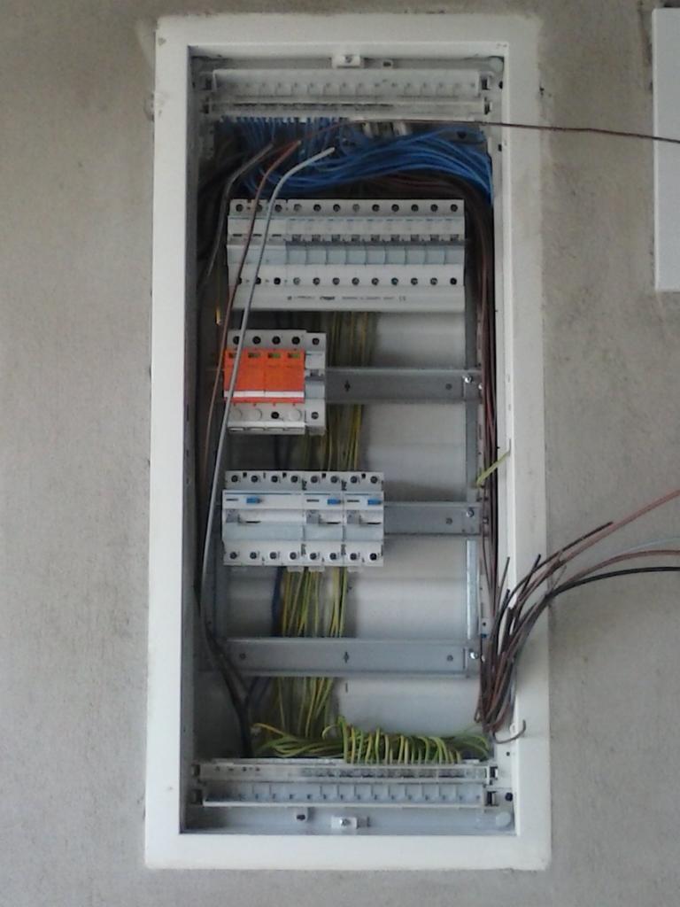 Otwarta skrzynka elektryczna z widocznymi bezpiecznikami, przewodami w różnych kolorach i metalowymi szynami montażowymi, osadzona w ścianie o surowym wykończeniu.