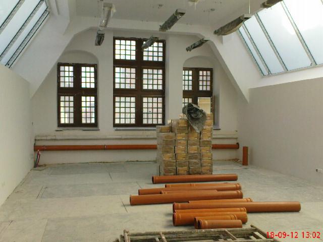 Wnętrze poddasza z oknami dachowymi i klasycznymi, przygotowane do prac instalacyjnych: widoczne pomarańczowe rury kanalizacyjne i paleta cegieł na posadzce.