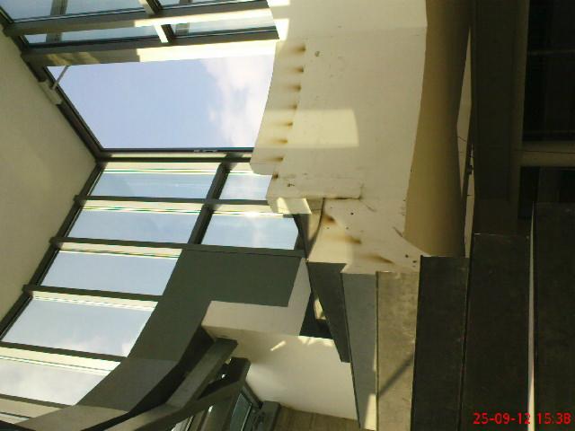 Fragment konstrukcji szklanego dachu z elementami stalowymi i betonowymi, widoczne ślady spawania na betonowym słupie, ujęcie z dołu.