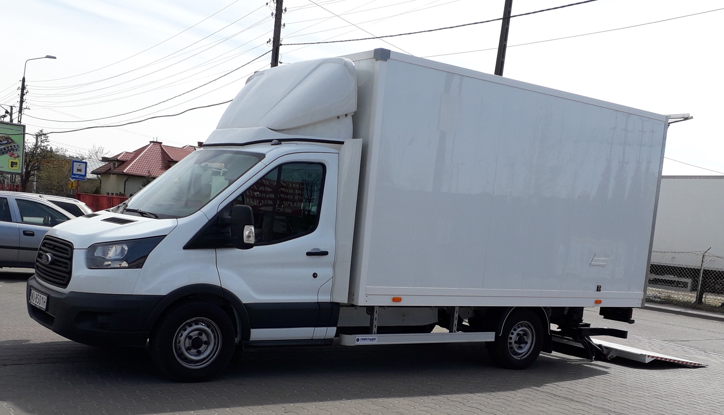 Biały samochód dostawczy z windą załadowczą na chodniku w słoneczny dzień.