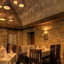 Eleganckie wnętrze restauracji z kamienną ścianą, drewnianymi krzesłami i stołami nakrytymi białymi obrusami. Oświetlenie punktowe z ażurowymi kloszami.