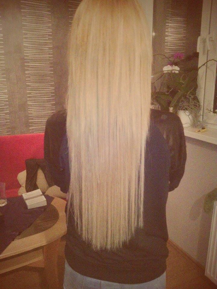 Długie, proste, blond włosy kobiety, widziane od tyłu. W tle widoczny fragment pokoju z kwiatami na parapecie i elementami wyposażenia.