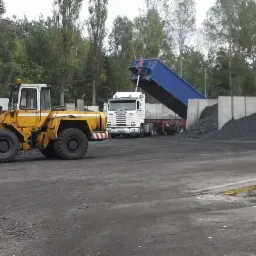 Żółta ładowarka kołowa i biała ciężarówka z niebieską naczepą wyładowująca węgiel na składzie, betonowe ściany oporowe w tle.