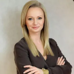 Portret kobiety w brązowej bluzce z długimi blond włosami i złotą bransoletką na tle jasnej ściany, pozującej z założonymi rękami.