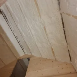 Poddasze z drewnianą konstrukcją pokryte warstwą izolacyjną z pianki, widoczna drabina oparta o ścianę.