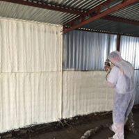 Pracownik w kombinezonie ochronnym aplikuje izolację natryskową na ściany budynku z blachy falistej, widoczne nierównomierne pokrycie i struktura piany.