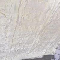 Świeżo nałożona warstwa izolacji na poddaszu, widoczne nierówności i tekstura piany, jasny odcień, fragment muru w rogu kadru.