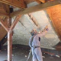 Pracownik w kombinezonie ochronnym aplikuje izolację natryskową na poddaszu z widoczną konstrukcją drewnianą. Zastosowana izolacja pokrywa skośną powierzchnię dachu, tworząc jednolitą warstwę.