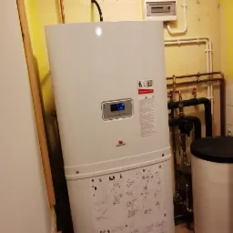 Biały piec gazowy z wyświetlaczem, zainstalowany w pomieszczeniu w Chwarstnicy, widoczne rury i grzejnik.