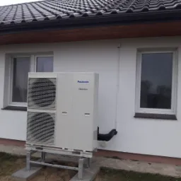 Biała pompa ciepła Panasonic Inverter, zamontowana na metalowej konstrukcji na betonowych podstawach, przy białej ścianie budynku z dwoma oknami i brązowym dachem z dachówki.