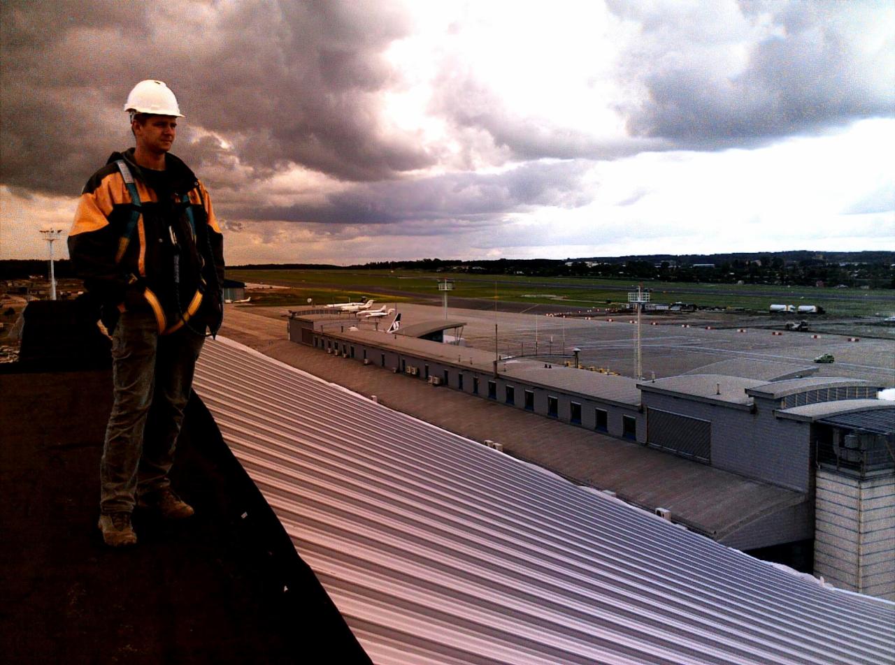 Mężczyzna w kasku i uprzęży stoi na blaszanym dachu budynku lotniska, obserwując pas startowy i samoloty na tle pochmurnego nieba.