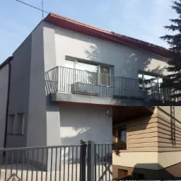 Szary dom z balkonem i metalową balustradą, widoczny fragment elewacji z poziomymi panelami w odcieniach beżu, ogrodzenie metalowe w pierwszym planie.