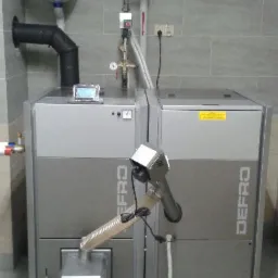 Szary, dwuczęściowy piec Defro z podajnikiem, rurami i zbiornikiem wyrównawczym w kotłowni z szarymi płytkami. Widoczne elementy instalacji i sterownik.