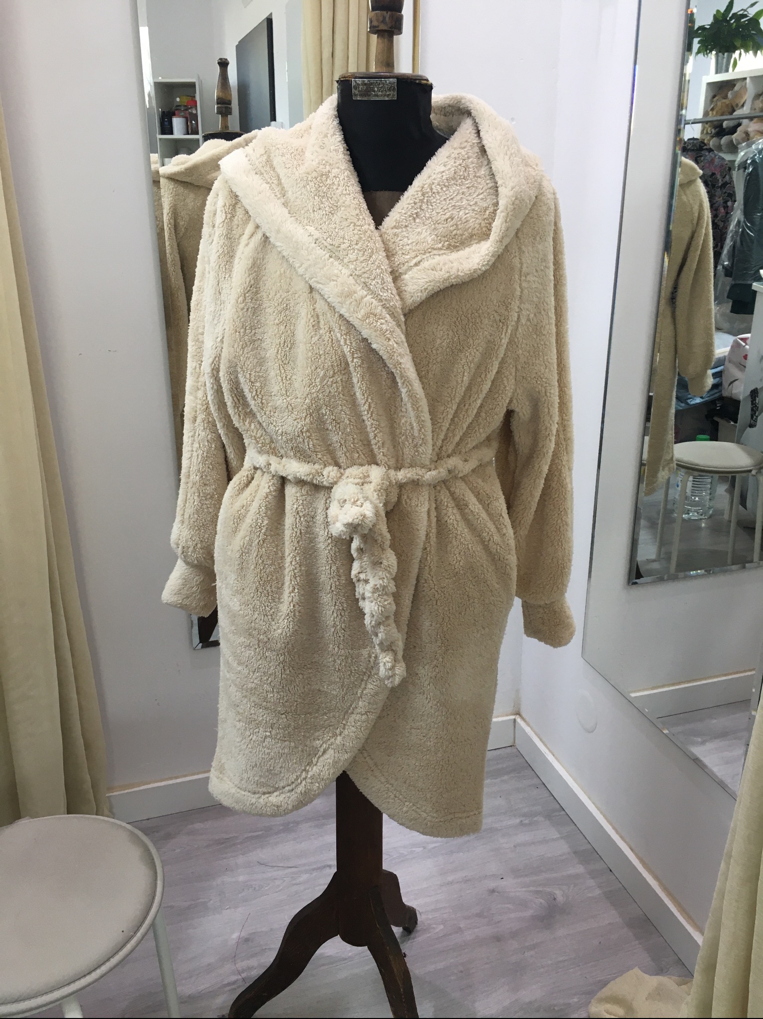 Kremowy, pluszowy szlafrok z kapturem i wiązaniem w pasie, prezentowany na drewnianym manekinie krawieckim w jasnym pomieszczeniu.