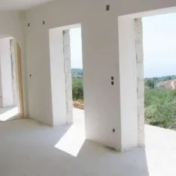 Wnętrze budynku po tynkowaniu: białe ściany, widoczne otwory okienne z kamiennymi obramowaniami i widok na zieleń za oknem. Gładkie, jasne powierzchnie ścian.