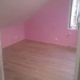 Pomalowane na różowo poddasze z drewnianą podłogą, okno w stylu gotyckim i widoczne gniazdka elektryczne.