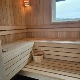 Wnętrze jasnej, drewnianej kabiny z ławkami, wiadrem i chochlą, oświetlone lampą ścienną, z widokiem na zewnątrz przez okno.