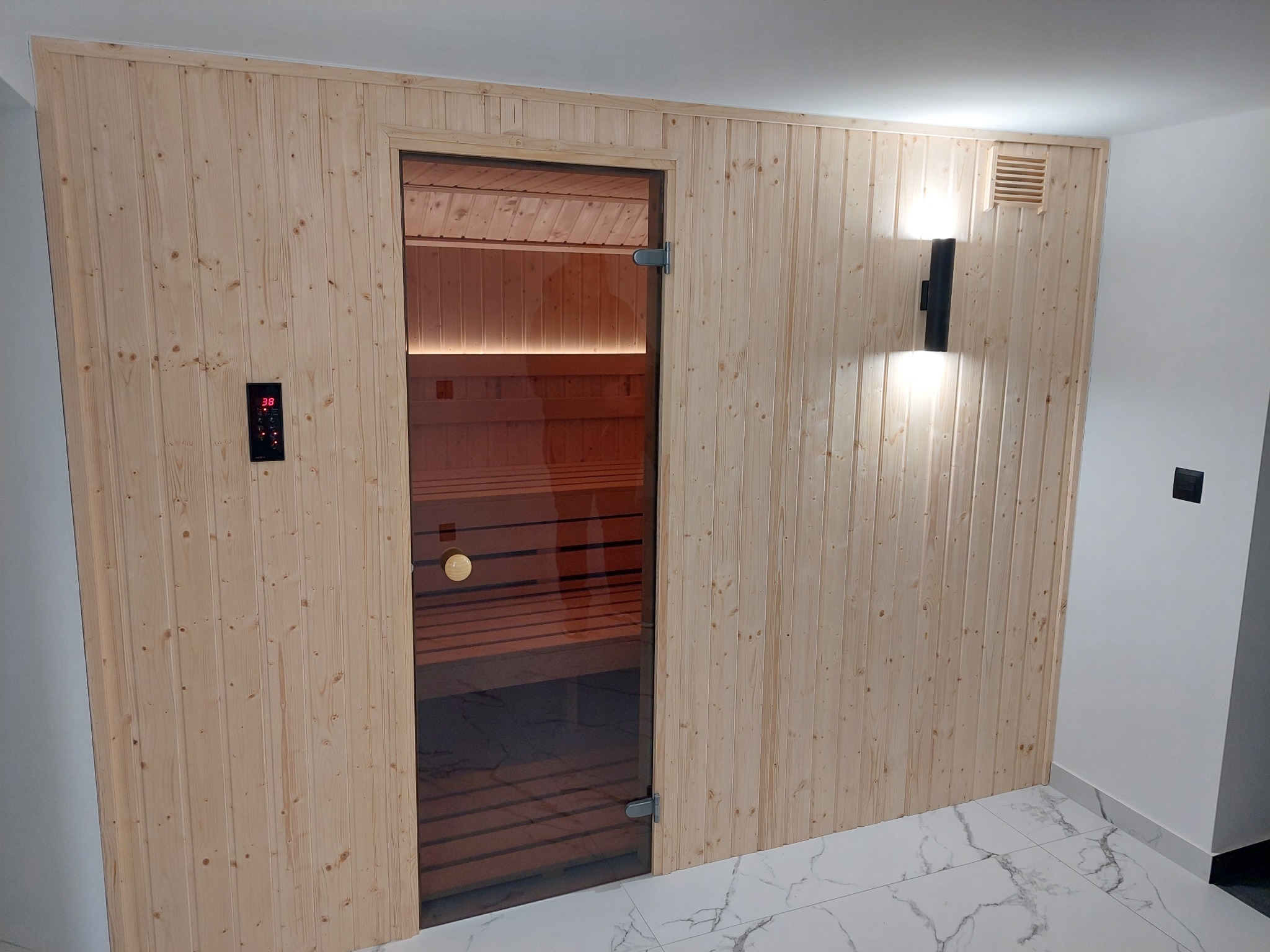Wnętrze wykończonej kabiny z przeszklonymi drzwiami, obudowanej jasnym drewnem z panelem sterowania temperaturą i oświetleniem, widoczna wentylacja oraz marmurowa posadzka.