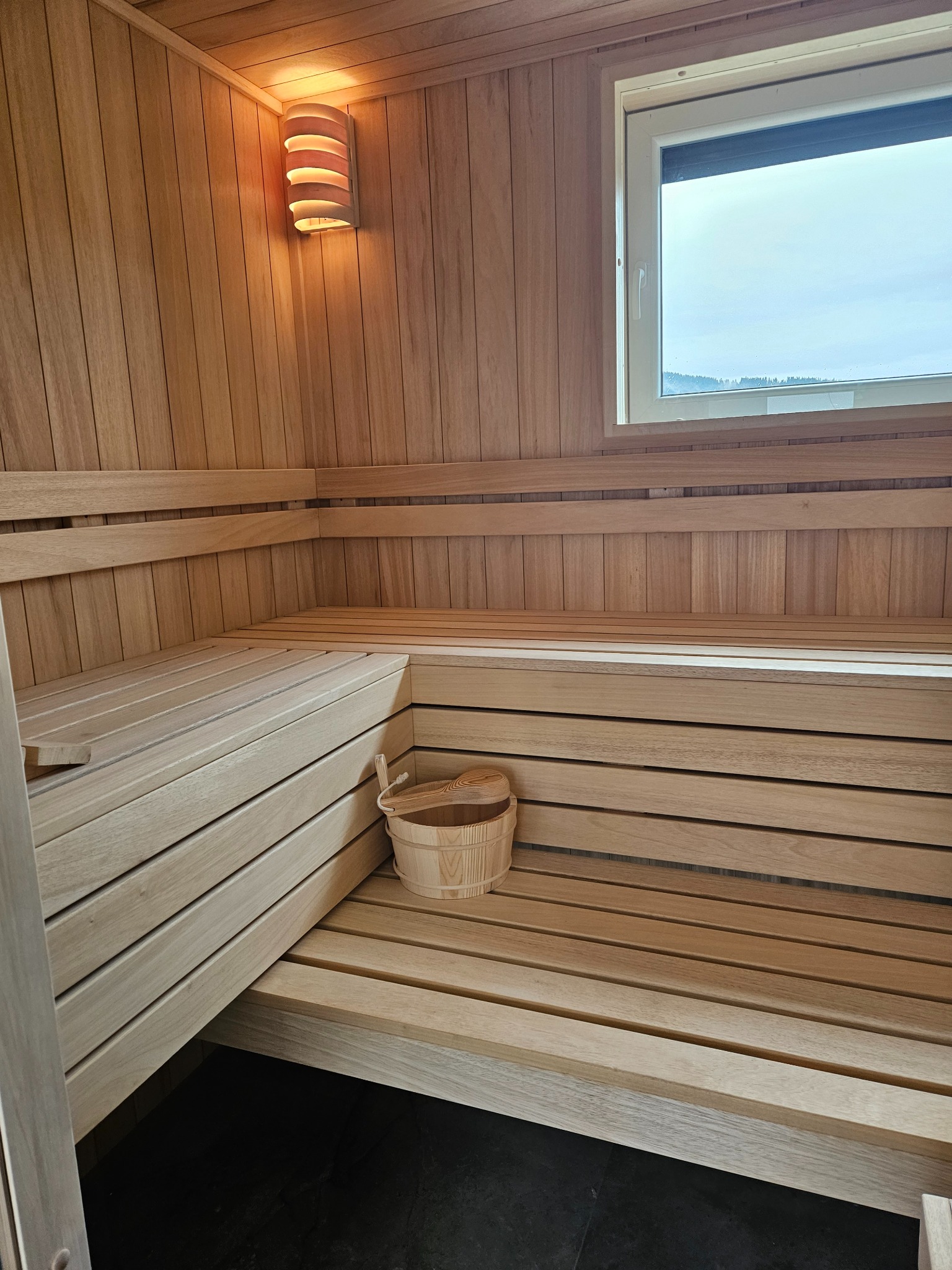Wnętrze jasnej, drewnianej kabiny z ławkami, wiadrem i chochlą, oświetlone lampą ścienną, z widokiem na zewnątrz przez okno.