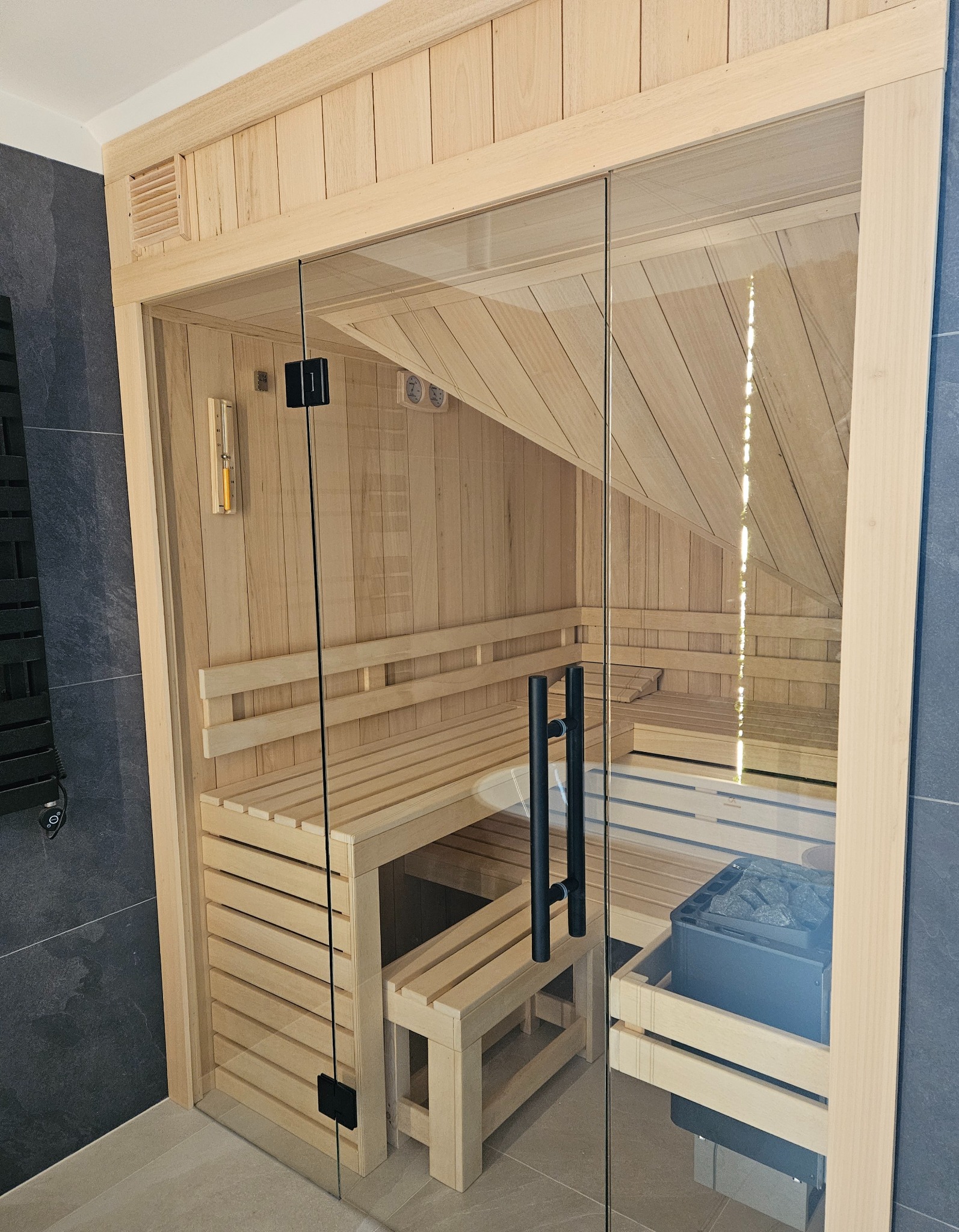 Nowoczesna kabina z jasnego drewna, z przeszklonymi drzwiami i czarnymi uchwytami, wewnątrz widoczne ławki, piec z kamieniami, termometr i higrometr.