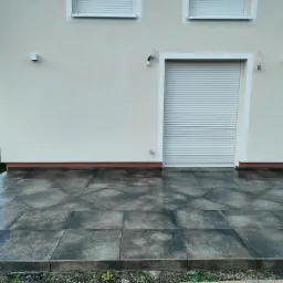 Wykończony taras z ciemnych, błyszczących płytek ceramicznych ułożonych przed budynkiem z roletami zewnętrznymi, widoczne niedociągnięcia w postaci roślinności przy krawędziach tarasu.