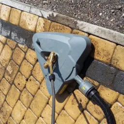 Czyszczenie kostki brukowej za pomocą myjki ciśnieniowej; widoczne różnice w kolorze kostki przed i po czyszczeniu, zarys krawężnika w tle.