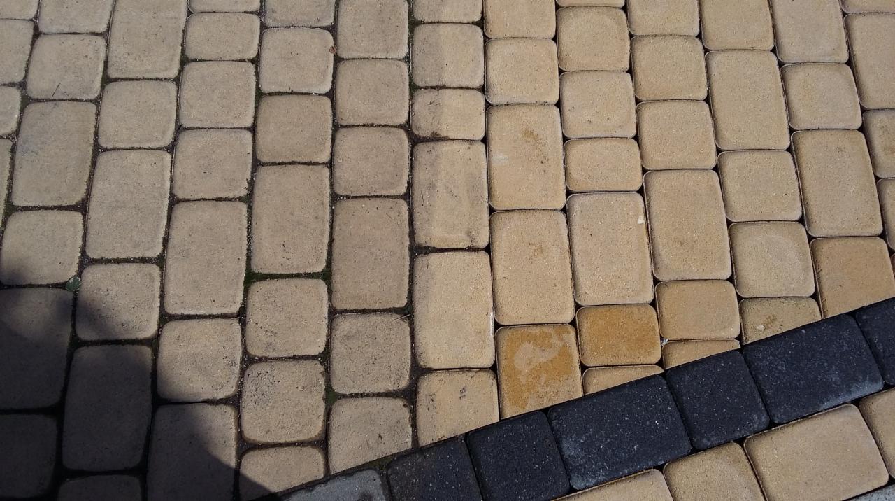 Fragment chodnika z kostki brukowej, część wyczyszczona, część zabrudzona, widoczne ciemne obrzeże.