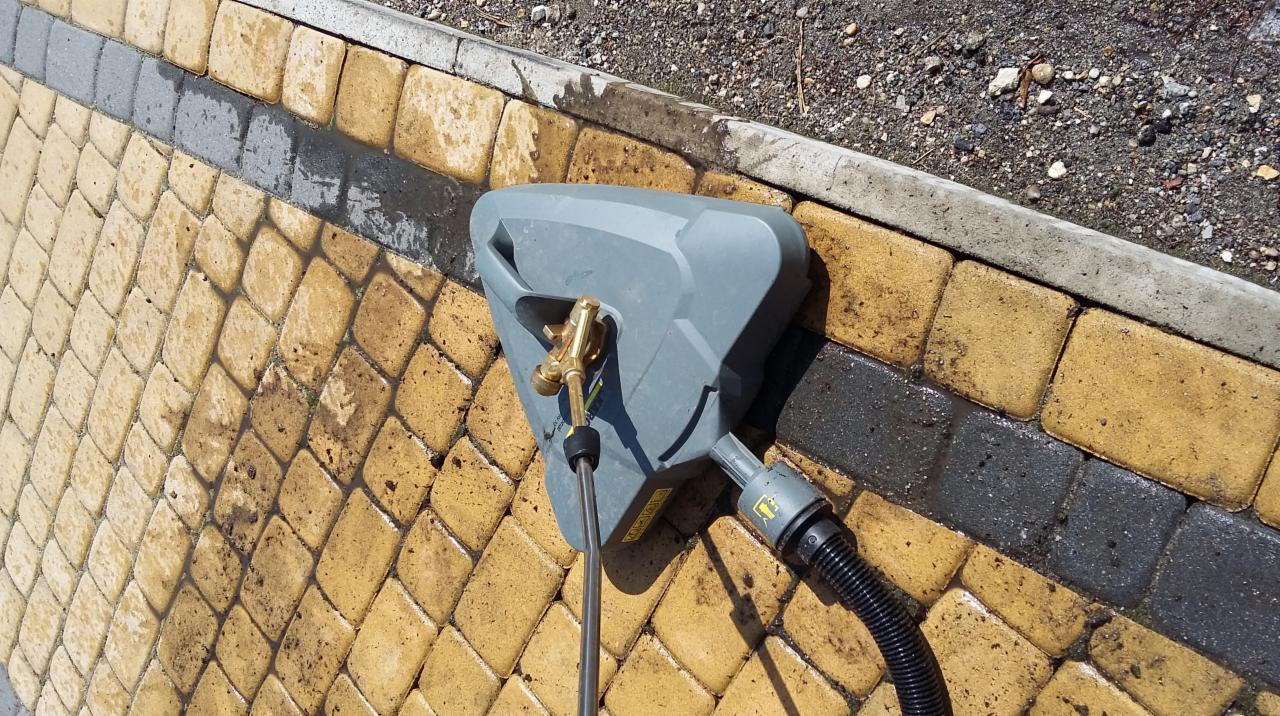 Czyszczenie kostki brukowej za pomocą myjki ciśnieniowej; widoczne różnice w kolorze kostki przed i po czyszczeniu, zarys krawężnika w tle.