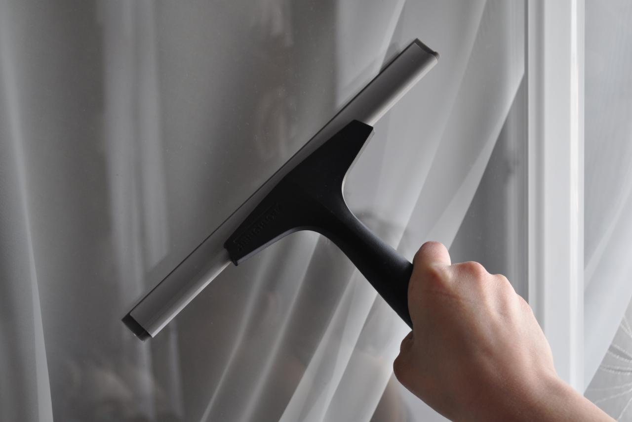Dłoń trzyma czarną myjkę do okien z gumową listwą na tle czystej szyby okiennej i białej firanki.