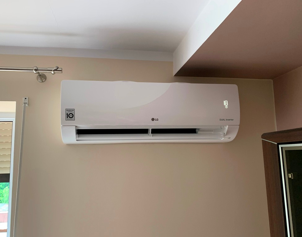 Biała jednostka klimatyzacji LG Dual Inverter zamontowana na beżowej ścianie w pokoju, widoczny fragment okna z żaluzjami i karnisz.
