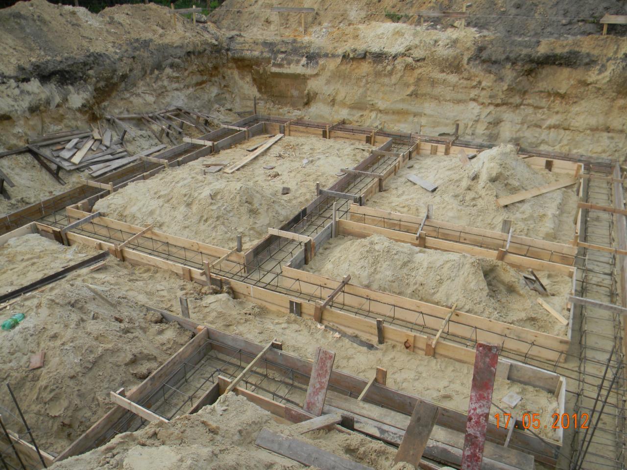 Przygotowanie fundamentów pod dom murowany: szalunek zbrojony stalą, wykop ziemny z widocznymi warstwami gruntu.