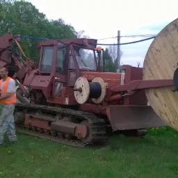 Czerwony traktor gąsienicowy z zamontowaną dużą szpulą kabla, w tle pracownicy i minikoparka, teren zielony.