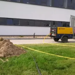 Żółty kompresor CompAir C38 na przyczepce podłączony do białego busa z otwartymi drzwiami, widoczny wykop w ziemi z żółtymi wężami na trawie przed budynkiem.