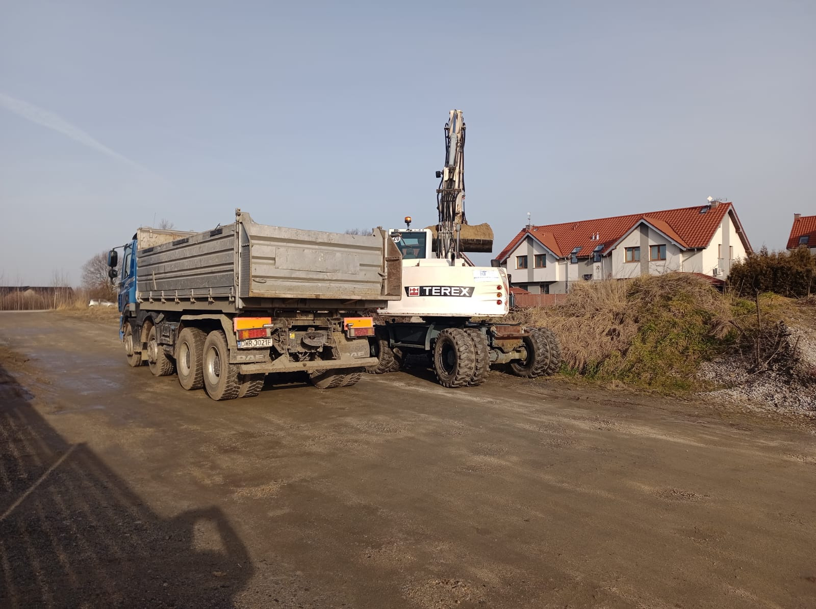 Ciężarówka i koparka Terex na placu budowy, w tle domy z czerwoną dachówką. Koparka w trakcie załadunku ziemi. Widoczne ślady opon na ziemi.