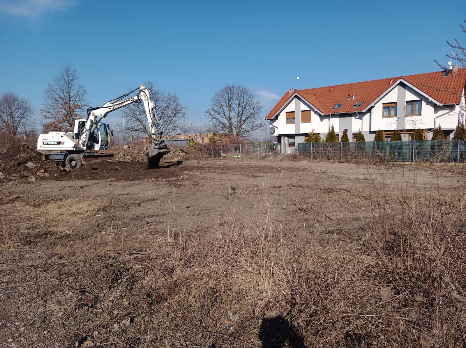 Biała koparka Terex na placu budowy, przygotowuje teren pod budowę domu jednorodzinnego w słoneczny dzień. Widoczne wykopane fragmenty ziemi.