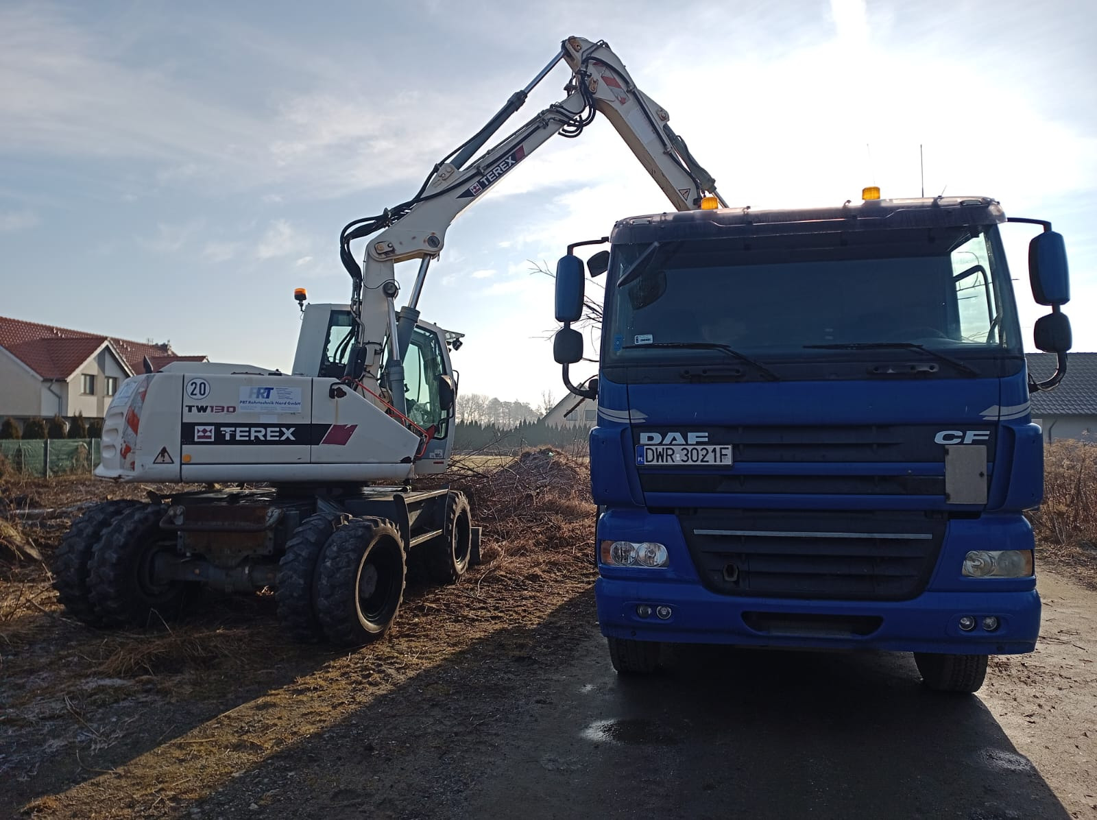 Biała koparka Terex TW130 i niebieska ciężarówka DAF CF na placu budowy, przygotowane do prac ziemnych. Słoneczny dzień, widoczne domy w tle.