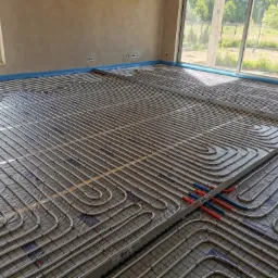 Układanie rur grzewczych na podkładzie w nowym budynku w Skierniewicach. Widoczne pętle rur, izolacja brzegowa i okna z widokiem na zieleń.