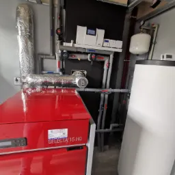 Kotłownia pelletowa po modernizacji: Czerwony piec na pellet marki Instal Service Selecta 15 HQ, biały zasobnik ciepłej wody użytkowej Kospel, rury izolowane termicznie, sterownik TECH...