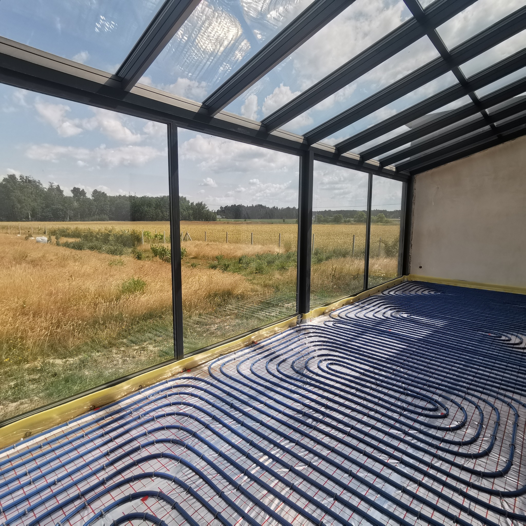 Niebieskie rury ogrzewania podłogowego ułożone spiralnie na folii aluminiowej z czerwoną siatką w przeszklonej werandzie z widokiem na pole zboża i błękitne niebo.