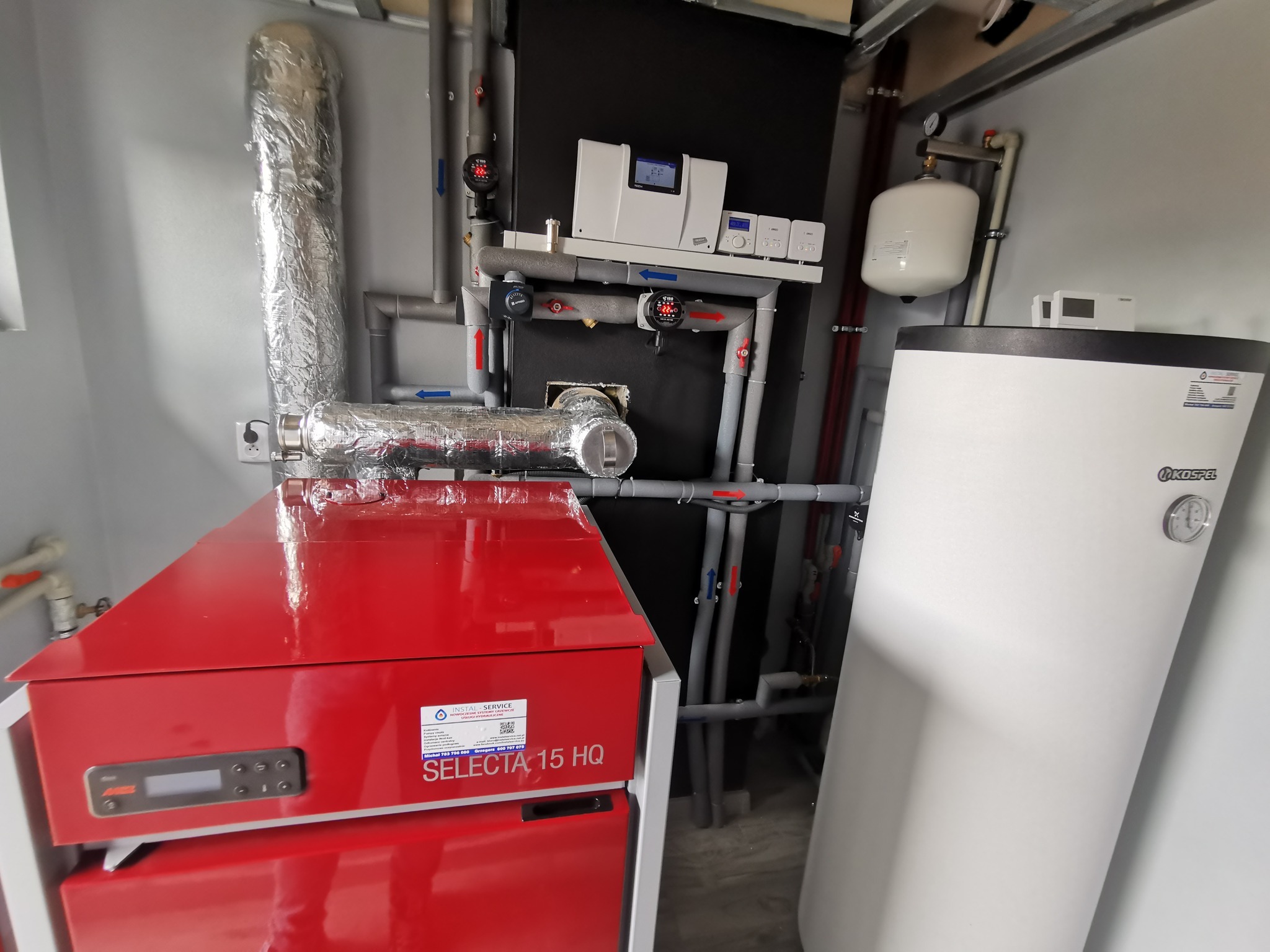 Kotłownia pelletowa po modernizacji: Czerwony piec na pellet marki Instal Service Selecta 15 HQ, biały zasobnik ciepłej wody użytkowej Kospel, rury izolowane termicznie, sterownik TECH...
