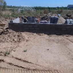 Budowa fundamentów z bloczków betonowych na piaszczystym terenie, widoczna betoniarka i ułożone bloczki, w tle konstrukcja szkieletowa budynku.