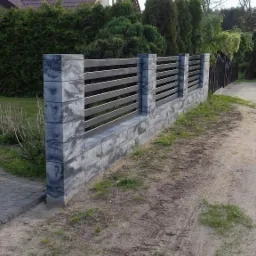 Nowoczesne ogrodzenie z betonowych bloczków w szaro-białej kolorystyce, z poziomymi metalowymi przęsłami, na tle zieleni i domu z brązowym dachem.