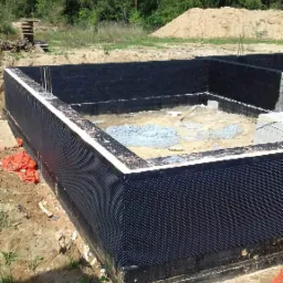 Fundamenty domu w trakcie budowy, zabezpieczone czarną folią kubełkową i izolacją, widoczne zbrojenia, w tle drzewa i usypana ziemia.