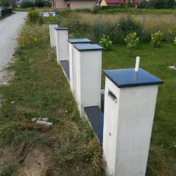 Rząd betonowych słupków z czarnymi daszkami, prawdopodobnie tworzących ogrodzenie lub skrzynki pocztowe, na tle wiejskiego krajobrazu z domami i zielenią.
